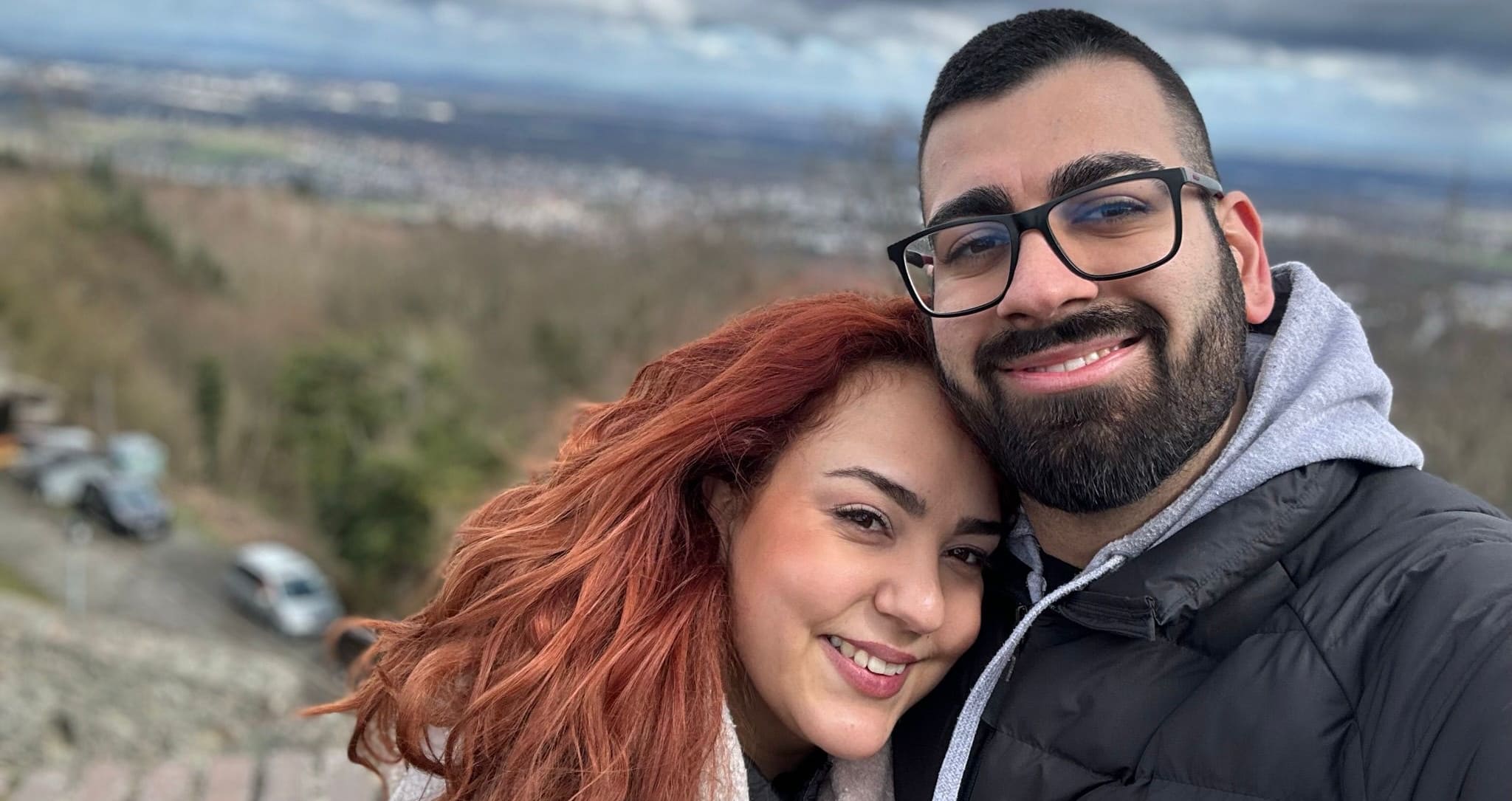 Margarita und Nikos, zwei junge griechische Menschen sind im Vordergrund des Bildes zu sehen, sie lächeln in die Kamera. Im Hintergrund, verschwommen, ein Dorf in einem Tal