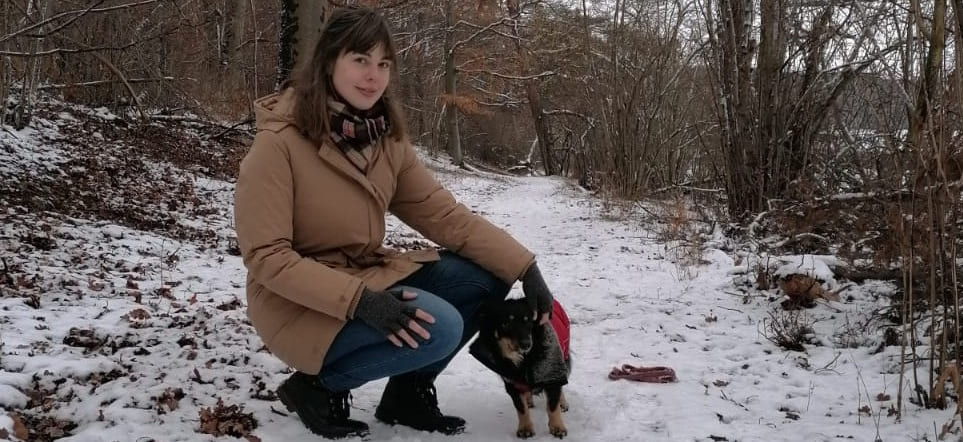 Mona lächelt in die Kamera, sie ist draußen, es ist Winter. Auf einem verschneiten Pfad, am Rande eines Waldstücks. Oh und ihr Hund Aluna ist auch dabei!